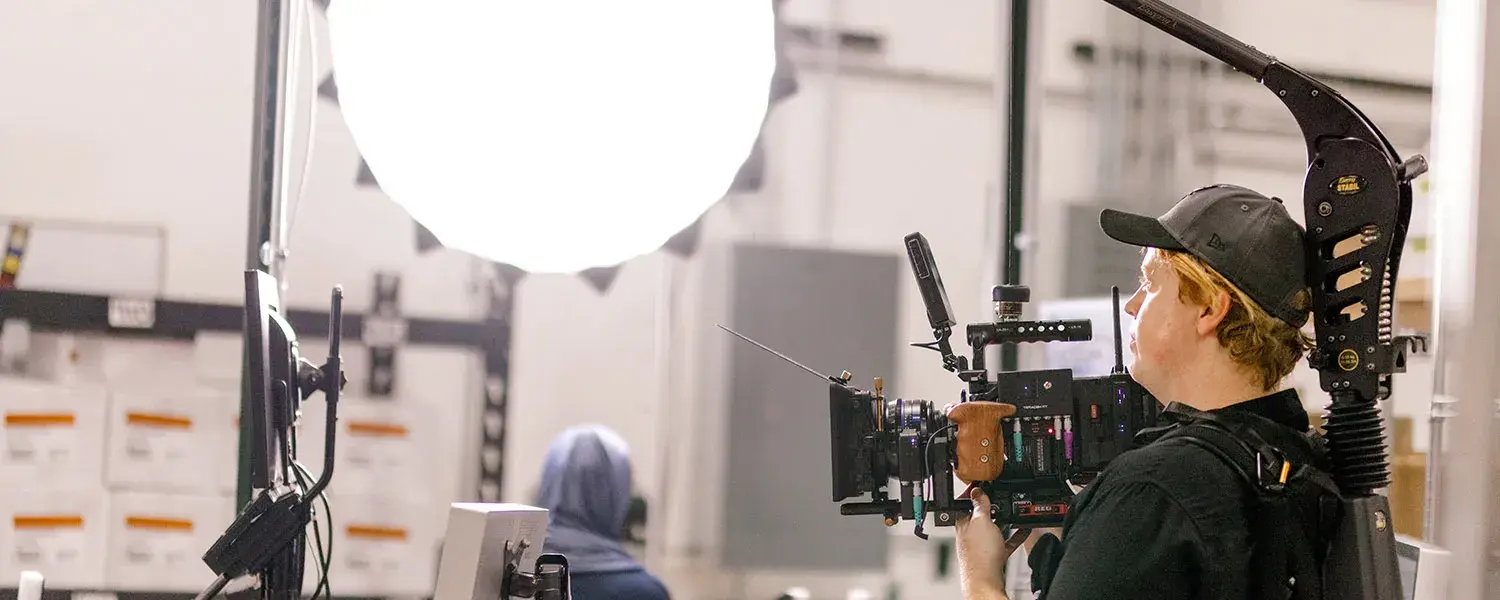 A man holds a large camera rig while filming on a video set.