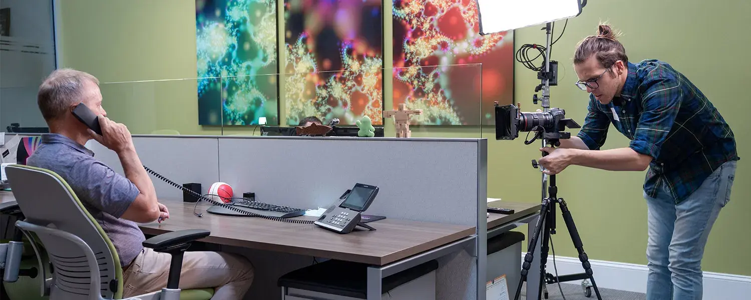 A man films a person taking a phone call at their office desk.